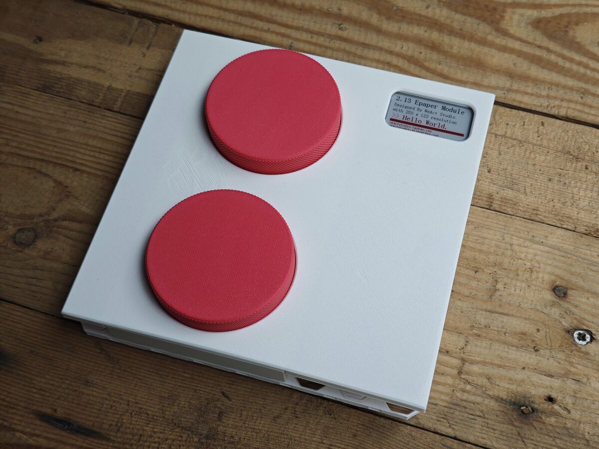 Deux gros boutons rotatifs coral sur une tuile blanche imprimée, étiquette e-paper, posés sur une table en bois
